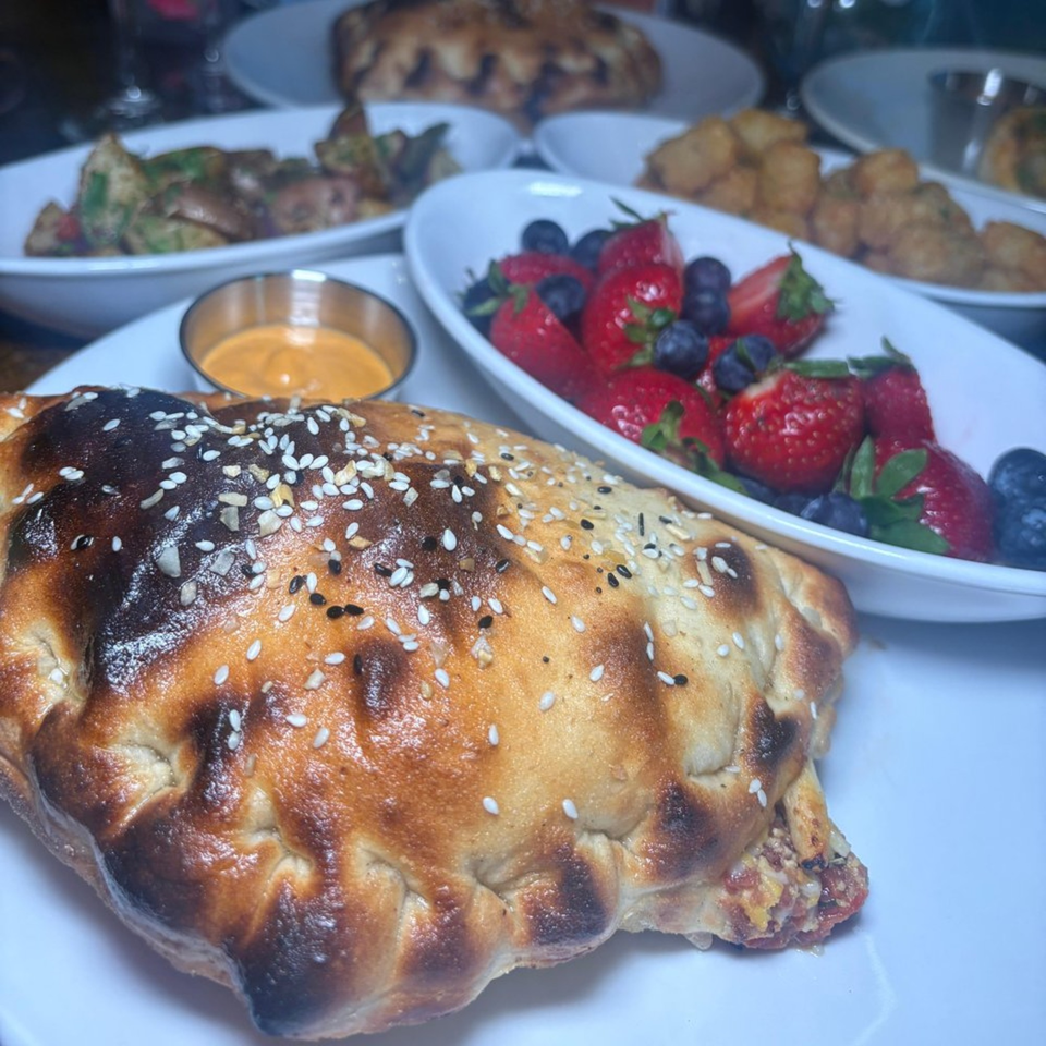 Calzone with sesame seeds, dipping sauce, and bowls of fruit and other dishes on a table.