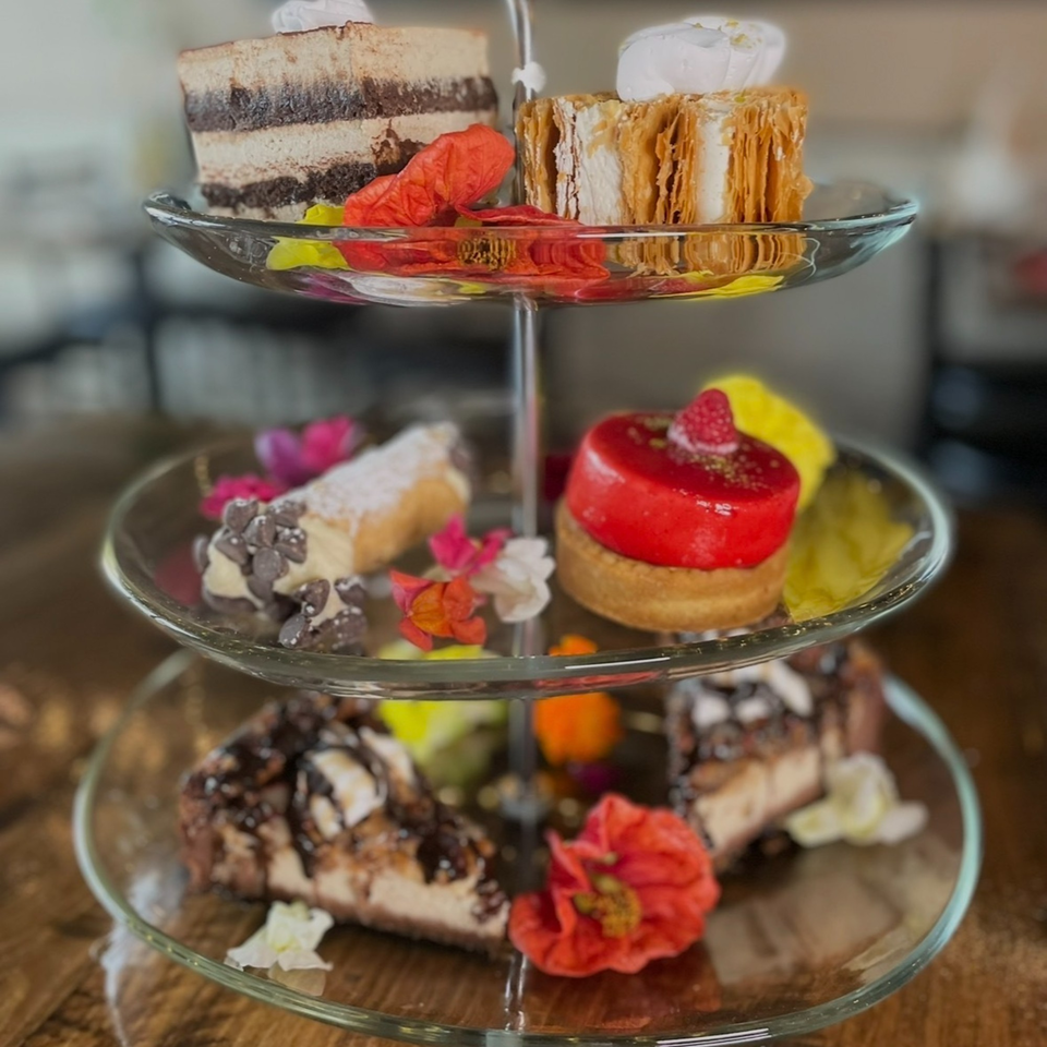 A three-tiered dessert stand filled with various pastries, including cakes, cannolis, and tarts, adorned with flowers.