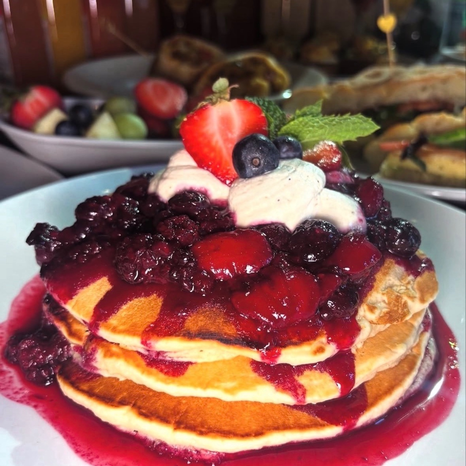 Stack of pancakes topped with berry compote, whipped cream, strawberries, blueberries, and mint on a plate.