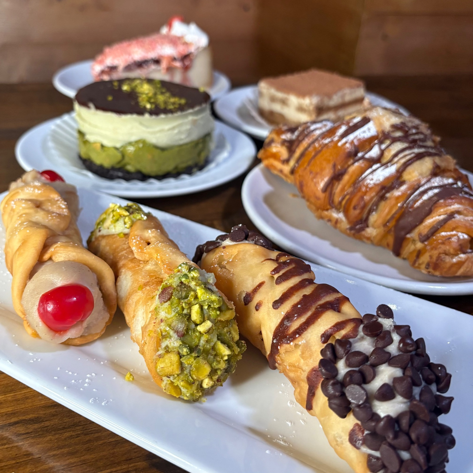 Assorted Italian pastries, including cream-filled cones, chocolate-drizzled pastries, cannoli, and layered cakes, arranged on white plates.