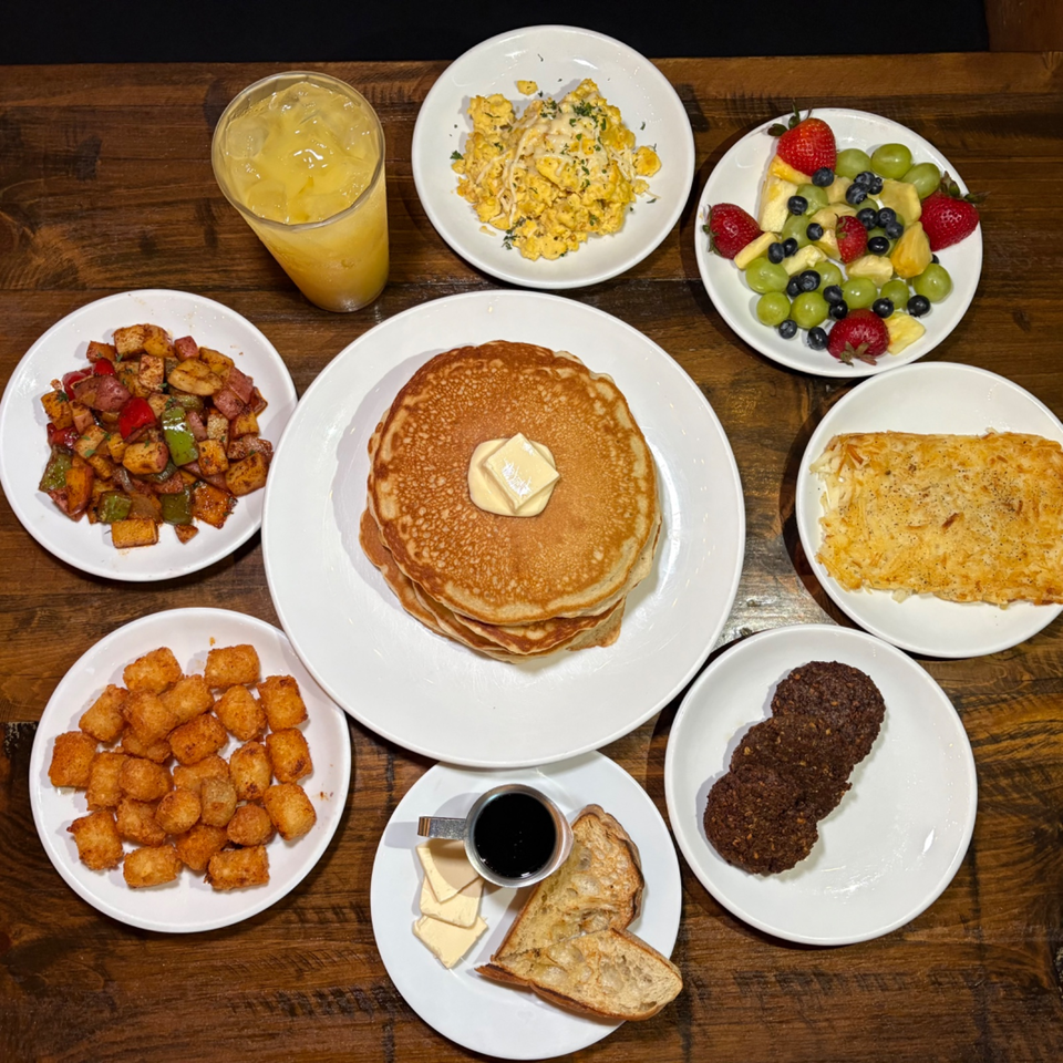 Overhead breakfast spread with pancakes, scrambled eggs, fruit, potatoes, hash browns, sausage patties, toast with syrup, and orange juice.