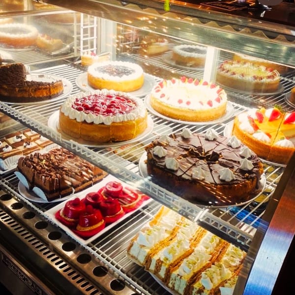 A brightly lit display case filled with various vegan cakes and pastries, including cheesecakes and chocolate cakes.