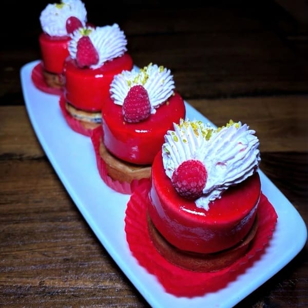 Four raspberry mini cakes with red glaze, whipped cream, and fresh raspberries on a white platter.