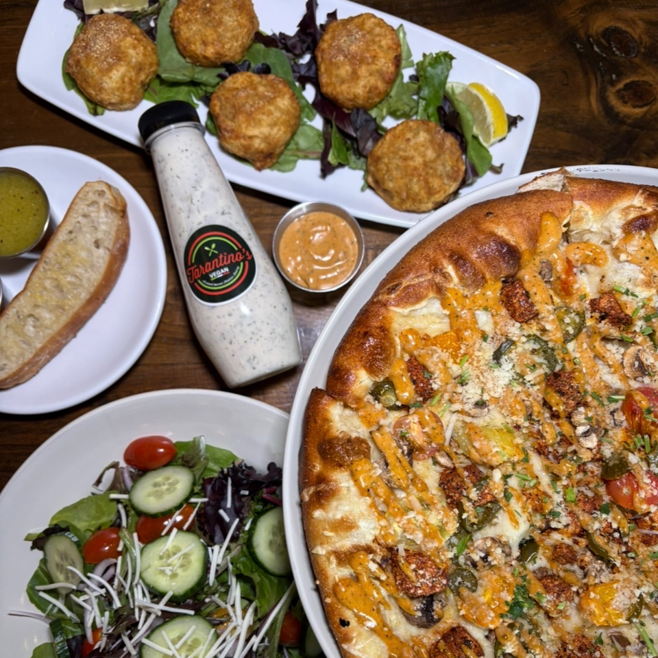 Assorted vegan dishes on a wooden table, including falafel, salad, dipping sauces, bread, and a loaded pizza.