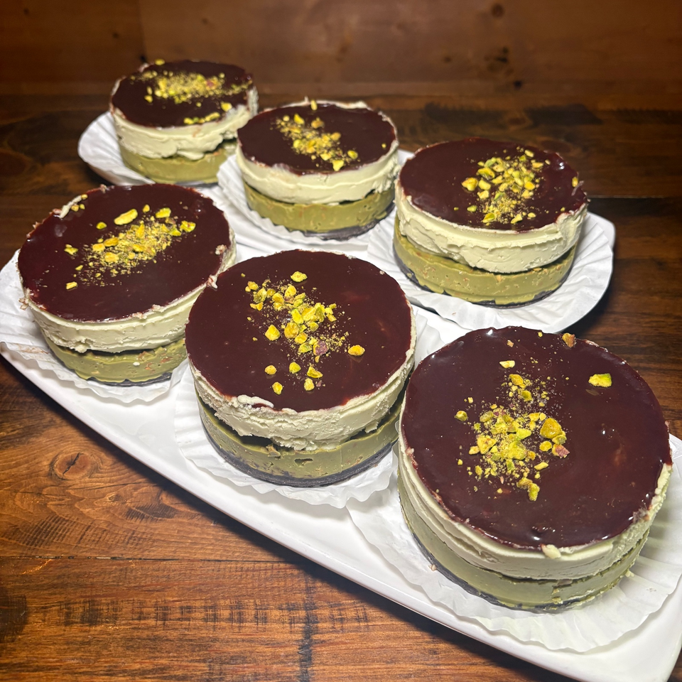 Six pistachio desserts layered with cream and topped with chocolate glaze and crushed pistachios on a white platter.