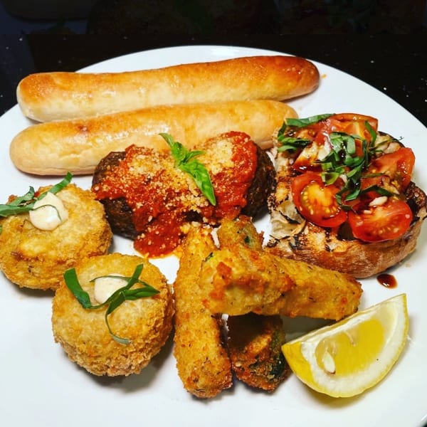 A white plate holds two breadsticks, a vegan meatball, fried appetizers, and bruschetta with a lemon wedge.