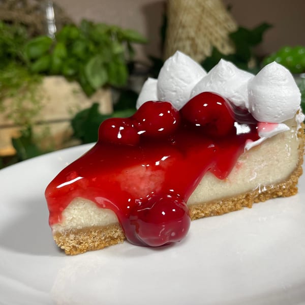 A slice of vegan cheesecake with a graham cracker crust, topped with cherry glaze and whipped cream.