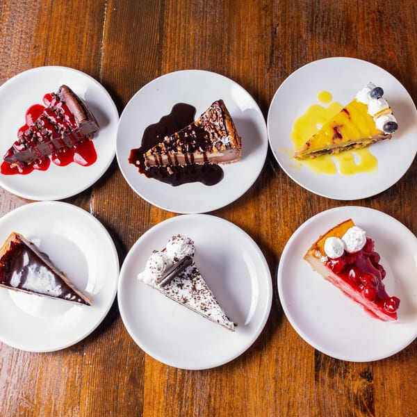 Six slices of vegan cheesecake with various toppings on white plates on a wooden table.