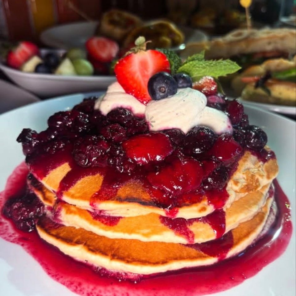 Stacked pancakes topped with berry compote, bananas, strawberries, blueberries, and cream on a plate.