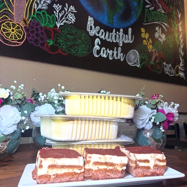 Three tiramisu slices on a white plate with stacked to-go containers and floral decorations.