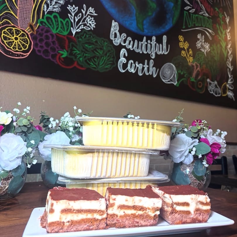 Three tiramisu slices on a white plate with stacked to-go containers and floral decorations.