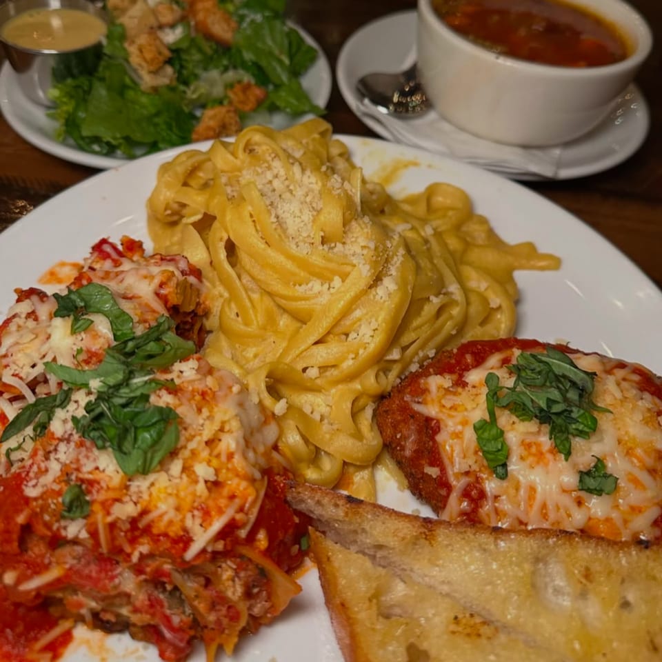 Vegan lasagna, fettuccine alfredo, chicken parmesan, garlic bread, salad, and soup on a table.