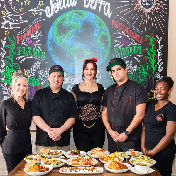 Five Tarantino's Vegan staff members stand proudly behind a table laden with various vegan dishes.