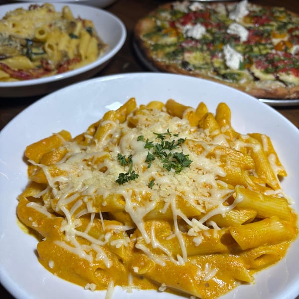 Creamy vegan penne pasta with melted cheese and parsley, with other dishes blurred in the background.
