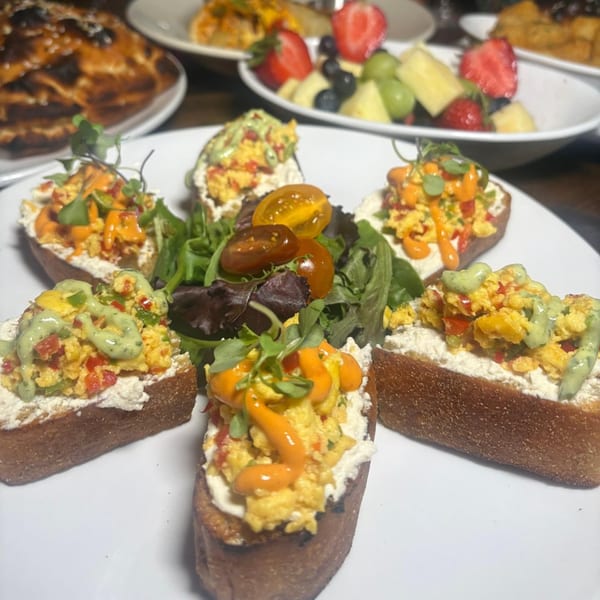 Six pieces of toast topped with scrambled eggs, sauces, and microgreens, with fruit in the background.