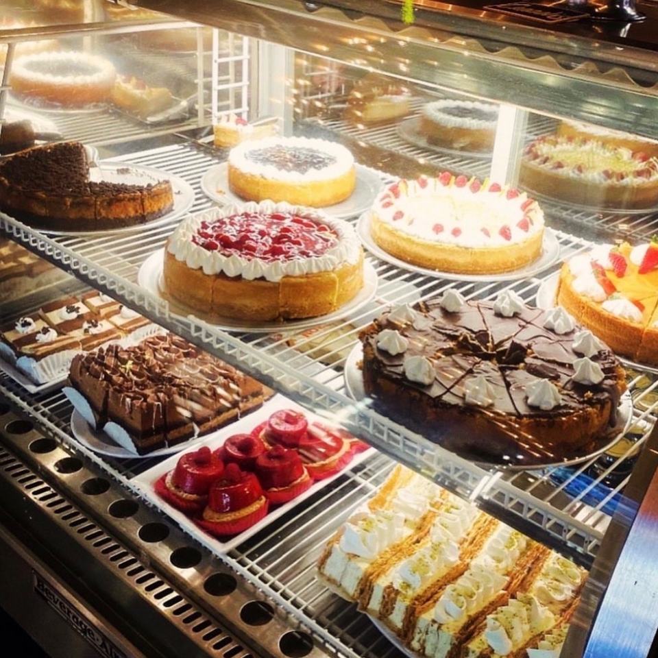 Glass display case filled with assorted cakes, cheesecakes, pastries, red fruit desserts, and cream-topped slices.