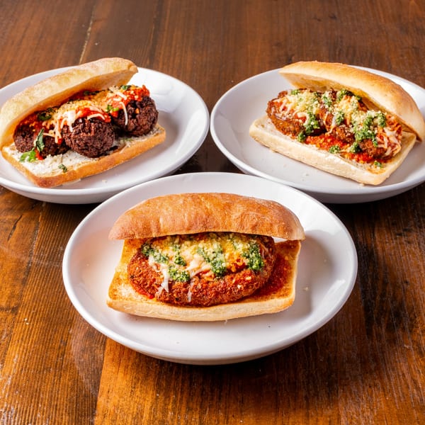 Three Tarantino's Vegan meatball and parmesan subs on white plates, on a wooden table.