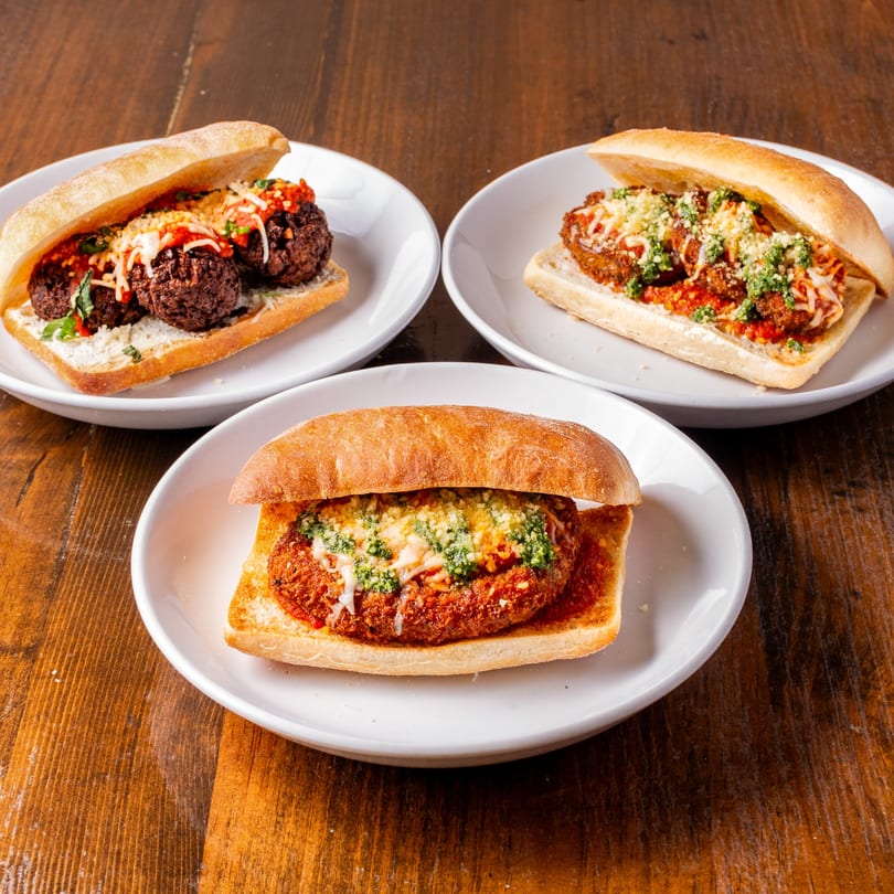Three Tarantino's Vegan meatball and parmesan subs on white plates, on a wooden table.