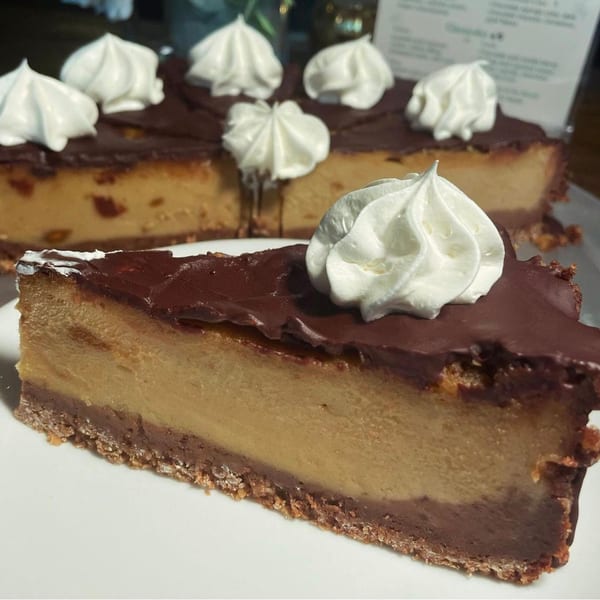 A slice of vegan cheesecake with chocolate topping and whipped cream, with the rest of the cake in the background.